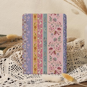 以下が含まれることがあります： ソフトピンクの背景と、紫、黄色、青の色合いのストライプが入った花柄のジャーナル。 ジャーナルは角が丸い長方形で、白いレースのテーブルクロスの上に置かれています。