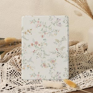 May include: A rectangular, off-white tablet case with a delicate floral pattern in pink, yellow, and purple. The design features trailing vines and small flowers. The case is set against a neutral background with decorative elements.
