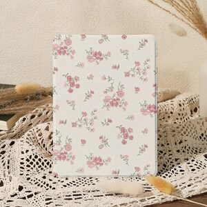 May include: A rectangular tablet case with a white background and a pattern of pink roses and green leaves. The case is sitting on a white lace tablecloth.