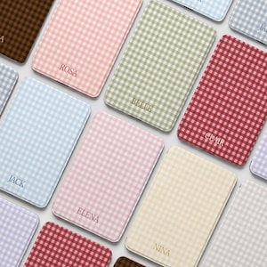 Puede incluir: Una colección de fundas rectangulares para teléfonos en varios colores pastel, cada una con un patrón de cuadros vichy. Las fundas están personalizadas con nombres en una fuente delicada, como "ROSA", "BELLE" y "CLAIR".