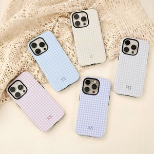 Puede incluir: Cinco fundas para teléfono con estampado de cuadros en colores pastel: azul claro, lavanda, crema y lila. Cada funda tiene un recorte negro para la cámara y unas iniciales en la esquina inferior. Las fundas están dispuestas sobre una superficie color crema.