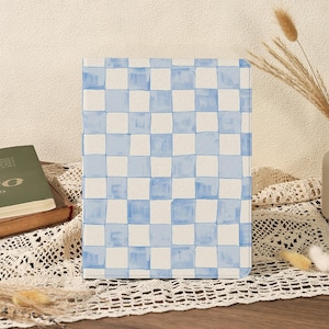 Può includere: Un taccuino rettangolare con un motivo a scacchi blu e bianco. Il design ad acquerello presenta quadrati azzurri su sfondo bianco. Il taccuino è esposto su una superficie di legno con un libro ed elementi decorativi sullo sfondo.