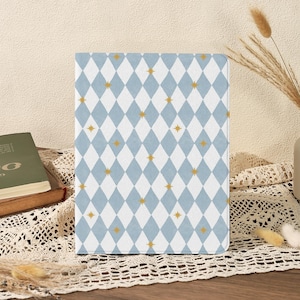 May include: A notebook cover with a light blue and white diamond pattern, interspersed with gold stars. The cover is displayed upright. The design is set against a wooden surface. The notebook is likely for writing.