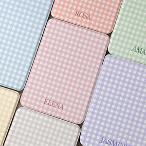 Op de afbeelding: Een collectie rechthoekige dienbladen met een gingham-patroon in diverse pastelkleuren, waaronder blauw, roze, groen, geel en grijs. Elk dienblad is gepersonaliseerd met een naam in een donker lettertype, zoals ROSA, ELENA en JASMINE.