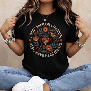 May include: A black t-shirt featuring a circular graphic with the text I HEAR MIGRANT CHILDREN ALSO HAVE HEARTBEATS. The design includes a central heart, a heartbeat line, orange flowers, blue leaves, and a small bird in a folk art style.