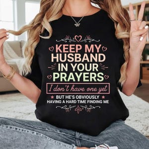 May include: Black t-shirt with the text "KEEP MY HUSBAND IN YOUR PRAYERS. I don't have one yet. But he's obviously having a hard time finding me." The text is in white, pink, and green. The shirt has a crew neck.
