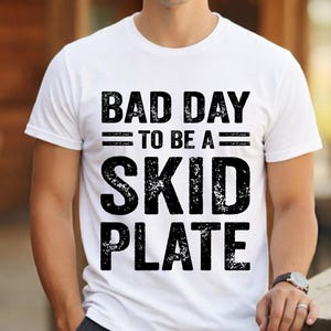May include: White crew neck t-shirt featuring the text "BAD DAY TO BE A SKID PLATE" in distressed black font. The shirt is being worn by a person.