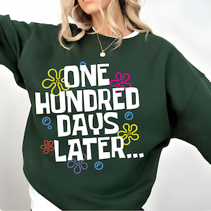 May include: Dark green sweatshirt with the text "ONE HUNDRED DAYS LATER..." in white. The design includes colorful flower and bubble graphics. The sweatshirt has a white collar.