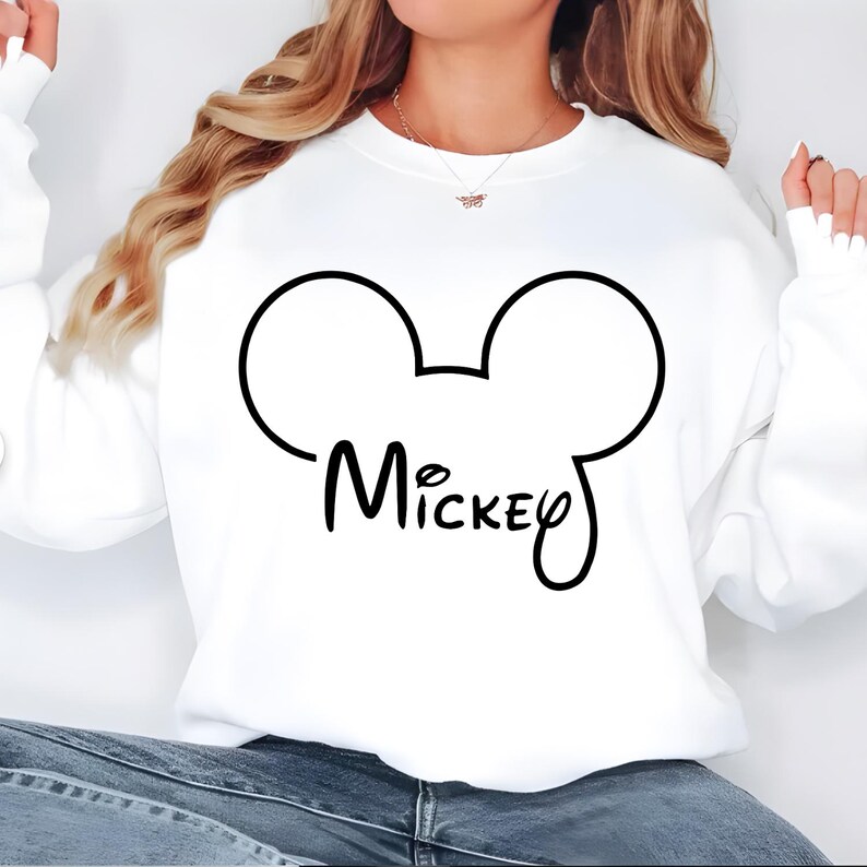 May include: White sweatshirt featuring a black outline of Mickey Mouse ears and the name "Mickey" in a script font. The sweatshirt is a casual style, suitable for everyday wear.