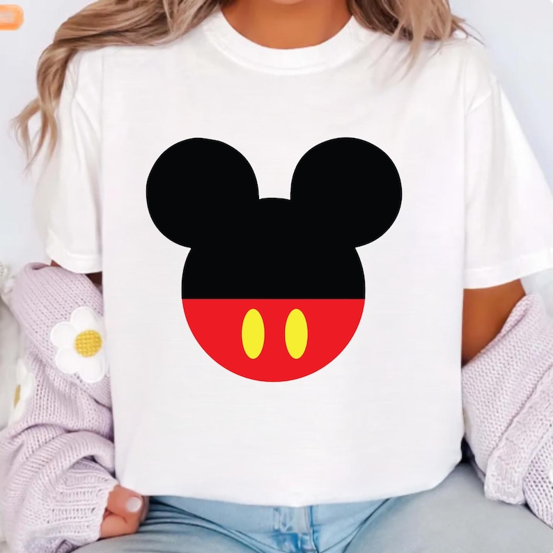 May include: White t-shirt featuring a large graphic of Mickey Mouse's face. The design includes black ears, a black upper face, a red lower face, and two yellow ovals. The shirt is a casual, everyday wear item.