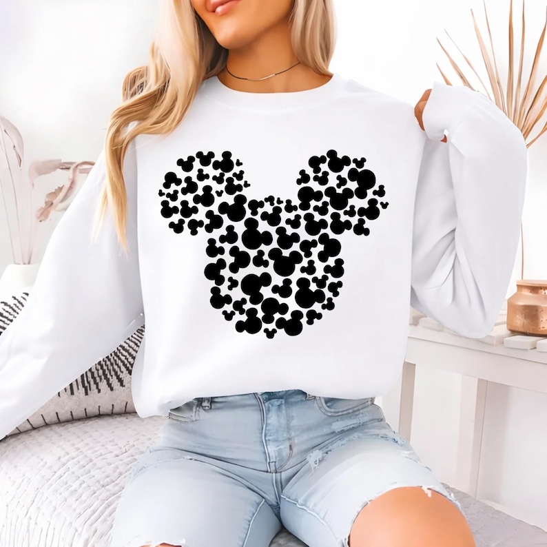 May include: White long-sleeve sweatshirt featuring a large black Mickey Mouse head design made of smaller Mickey Mouse silhouettes. The sweatshirt is a casual style, suitable for everyday wear.