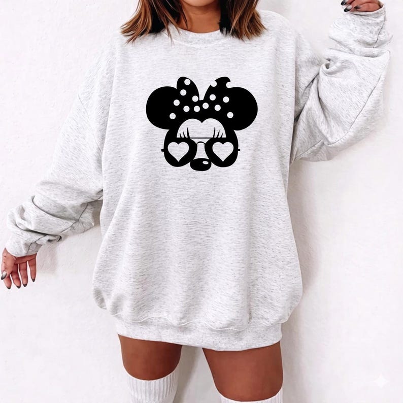 May include: A light gray sweatshirt featuring a black Minnie Mouse design. The design includes a polka-dot bow, heart-shaped glasses, and mouse ears. The sweatshirt has long sleeves and a crew neck.