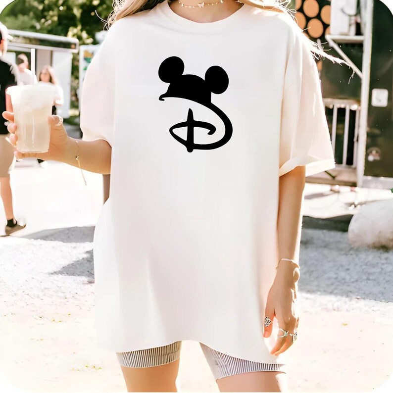 May include: Cream-colored t-shirt featuring a black Mickey Mouse logo. The oversized tee has short sleeves and a crew neck. The design is a classic Disney icon, perfect for fans of all ages.