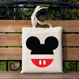 May include: A beige canvas tote bag featuring a black Mickey Mouse silhouette and a red mouth with two white dots. The bag has two beige handles and is displayed outdoors.