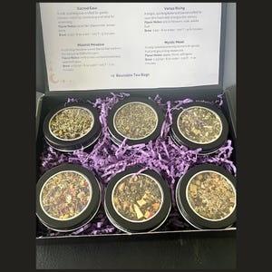 May include: A gift set of six tins of loose leaf tea, each with a different blend. The tins are black with clear lids, nestled in purple paper. A card lists the tea names and flavor notes. Includes two reusable tea bags.