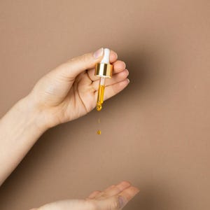 May include: A dropper with a white top and gold-colored metal is held over an open hand. A golden liquid is dripping from the dropper. The background is a solid, light brown color. The image is a close-up.