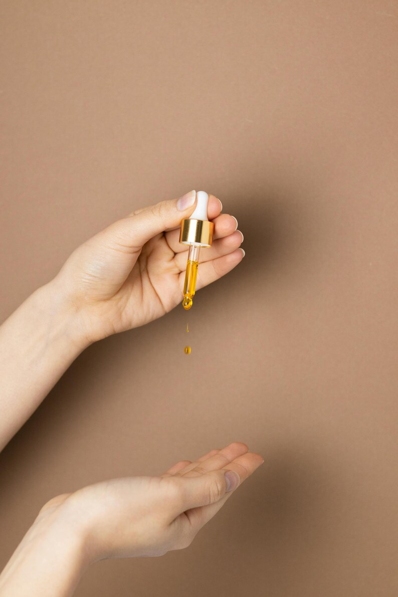 May include: A dropper with a white top and gold-colored metal is held over an open hand. A golden liquid is dripping from the dropper. The background is a solid, light brown color. The image is a close-up.