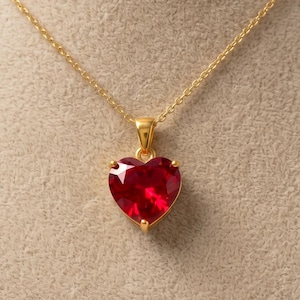 May include: A gold-colored necklace with a heart-shaped, red gemstone pendant. The pendant is a vibrant red color and is set in a gold-colored metal. The necklace is displayed on a mannequin.