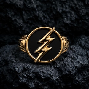 May include: A gold-toned ring featuring a circular face with a black inlay. Centered on the black background is a raised, metallic gold lightning bolt symbol. The band of the ring has a textured, sculpted design on the sides of the circular face.