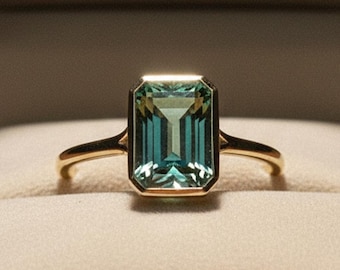 May include: A gold ring featuring a rectangular emerald-cut teal gemstone in a bezel setting, presented inside a dark brown jewelry box with the word STELLAR printed in gold lettering on the interior lid of the cream-colored velvet lining.