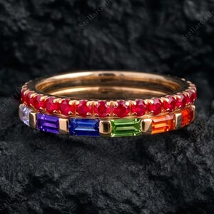 May include: A gold ring featuring two stacked bands. The top band is set with a row of small, round, vibrant red gemstones. The bottom band features a row of rectangular, baguette-cut gemstones in a rainbow sequence of purple, blue, green, and orange.