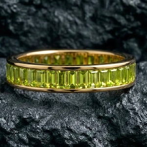 Puede incluir: Un anillo de oro con una banda de piedras preciosas rectangulares de color verde claro. El anillo se encuentra sobre un fondo oscuro y texturizado, resaltando el color vibrante de las piedras. El diseño del anillo es simple pero elegante, adecuado para el uso diario o ocasiones especiales.
