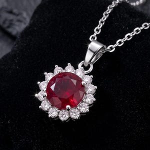 May include: A silver pendant necklace featuring a large, round, deep red gemstone at the center, surrounded by a halo of small, clear, sparkling stones. The pendant hangs from a delicate silver chain, creating a classic, elegant look.