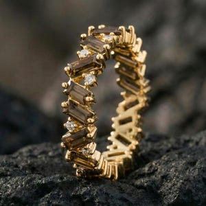 Puede incluir: Una alianza de oro con un patrón repetitivo de piedras de cuarzo ahumado rectangulares en talla baguette que se alternan con pequeños diamantes blancos redondos y transparentes, engastados en un marco de metal continuo y estructurado.
