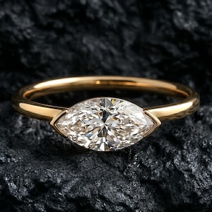 May include: A gold ring with a marquise-cut diamond. The diamond is set in a bezel setting, and the band is a simple, polished gold. The ring is displayed on a dark, textured surface.