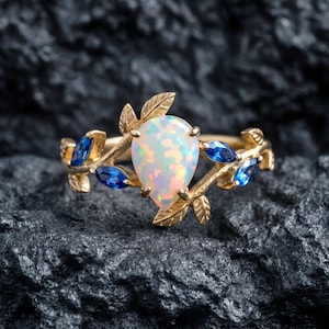 May include: A gold ring featuring a central pear-shaped opal gemstone surrounded by small marquise-cut blue stones. The band is designed with a textured leaf motif that branches out from the center, framing the iridescent opal and blue accents.