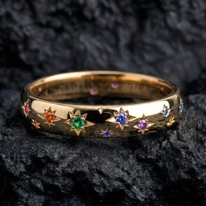 Könnte beinhalten: Ein goldener Bandring mit einer Reihe eingravierter Sternmotive, die jeweils mit einem kleinen farbigen Edelstein besetzt sind, darunter grüne, blaue, orange, rosa und violette Steine, die in einem himmlischen Muster auf der Außenseite angeordnet sind.