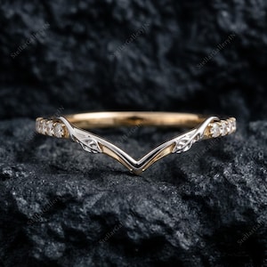 May include: A V-shaped ring crafted from gold, featuring a central point with engraved leaf-like detailing. The band is accented with small, clear round stones set along both sides, extending from the center toward the back of the ring.