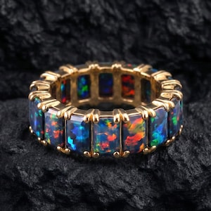 Op de afbeelding: Een gouden ring met een doorlopende rij rechthoekig geslepen zwarte opalen. Elke opaal vertoont levendige flitsen van rood, oranje, groen en blauw kleurenspel, stevig gezet in een gepolijst gouden metalen frame dat de gehele ring omringt.