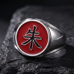 May include: A silver-toned metal ring featuring a circular face with a vibrant red enamel inlay. Centered on the red background is a black Japanese kanji character, specifically the symbol for vermilion or red, rendered in a bold, clean script.