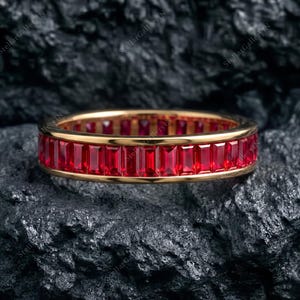 Puede incluir: Una alianza de oro con una hilera continua de piedras preciosas rojas de corte baguette en engaste de canal. El anillo tiene un acabado metálico pulido que contrasta con el color rojo intenso de las piedras, creando una forma circular definida.