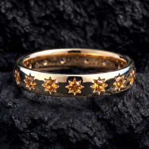 May include: A polished gold band ring featuring a series of evenly spaced, star-shaped settings. Each star setting contains a small, round, amber-colored gemstone. The ring has a smooth, reflective surface and a consistent width throughout the band.
