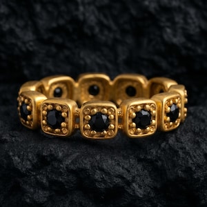 May include: A gold-colored ring with a repeating pattern of square black gemstones. The ring is set against a dark, textured background, highlighting the contrast between the gold and black elements. The ring's design features small, circular details around each gemstone.