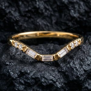 以下が含まれることがあります： ゴールドのシェブロン型リング。バンドの上部カーブに沿って、透明な長方形のバゲットカットストーンと小さなラウンドカットストーンが並んでいます。金属部分は光を反射する磨き上げられた仕上げになっています。