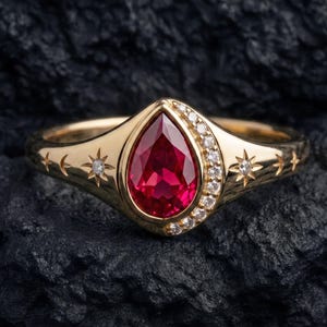 May include: A gold ring featuring a central pear-shaped red gemstone surrounded by a crescent of small white stones. The band is engraved with star and crescent moon motifs, with a small white stone set into the center of each star.