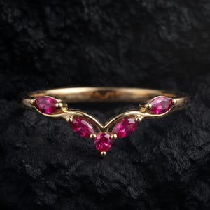 May include: A gold chevron-shaped ring featuring five marquise-cut vibrant red gemstones arranged in a V-pattern along the band. The ring has a polished metallic finish and a curved design that follows the contour of the finger, showcasing the stones.