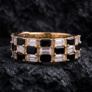 May include: A gold band ring featuring a checkerboard pattern of alternating baguette-cut clear white stones and rectangular black stones. The ring is constructed with a metal setting that holds the stones in a continuous, repeating grid design.