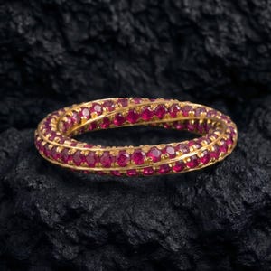 May include: A gold eternity ring featuring a continuous band of small, round, vibrant red rubies set in a twisted, interlocking design. The ring displays a polished metallic finish and a uniform arrangement of gemstones around the entire circumference.