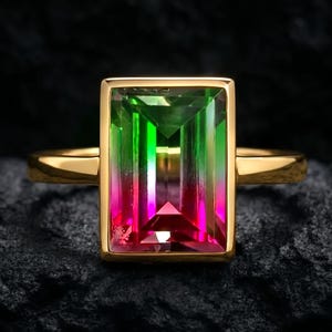 May include: A gold ring with a rectangular gemstone, displaying a gradient of green to pink hues. The gemstone is set in a gold bezel. The ring is presented on a dark, textured surface.