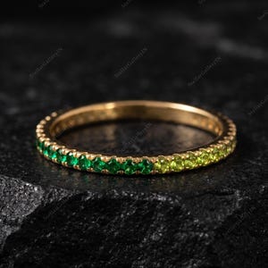 May include: A thin gold band ring featuring a continuous row of small, round-cut gemstones that transition in color from deep emerald green to a lighter, vibrant lime green, creating a gradient effect along the entire circumference of the ring.