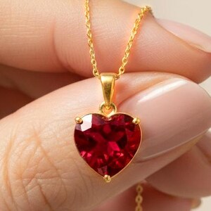 May include: A gold-colored necklace featuring a heart-shaped, deep red gemstone pendant. The pendant is held in a four-prong setting and hangs from a delicate chain. The necklace is a piece of jewelry.