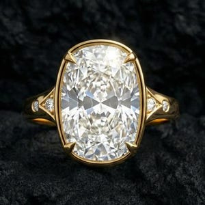 May include: A gold engagement ring with a large, oval-cut diamond as the centerpiece. The ring has a delicate band with smaller diamonds on each side of the main stone. The ring is set against a dark, textured background.