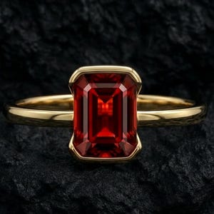 May include: A gold ring with a large, rectangular, deep red gemstone. The gemstone is set in a gold bezel, and the band is a simple, polished gold. The ring is displayed on a dark, textured surface.