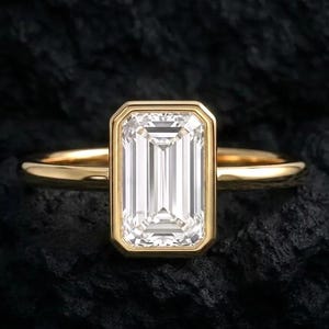 May include: A gold ring with an emerald-cut diamond. The diamond is set in a bezel setting and has a rectangular shape. The band is a simple, polished gold. The ring is displayed on a dark, textured surface.