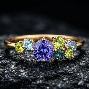 Peut inclure: Une bague en or ornée d'une grosse pierre violette ronde au centre, entourée de petites pierres colorées vertes, bleues et turquoise. La bague est posée sur une surface sombre et texturée.