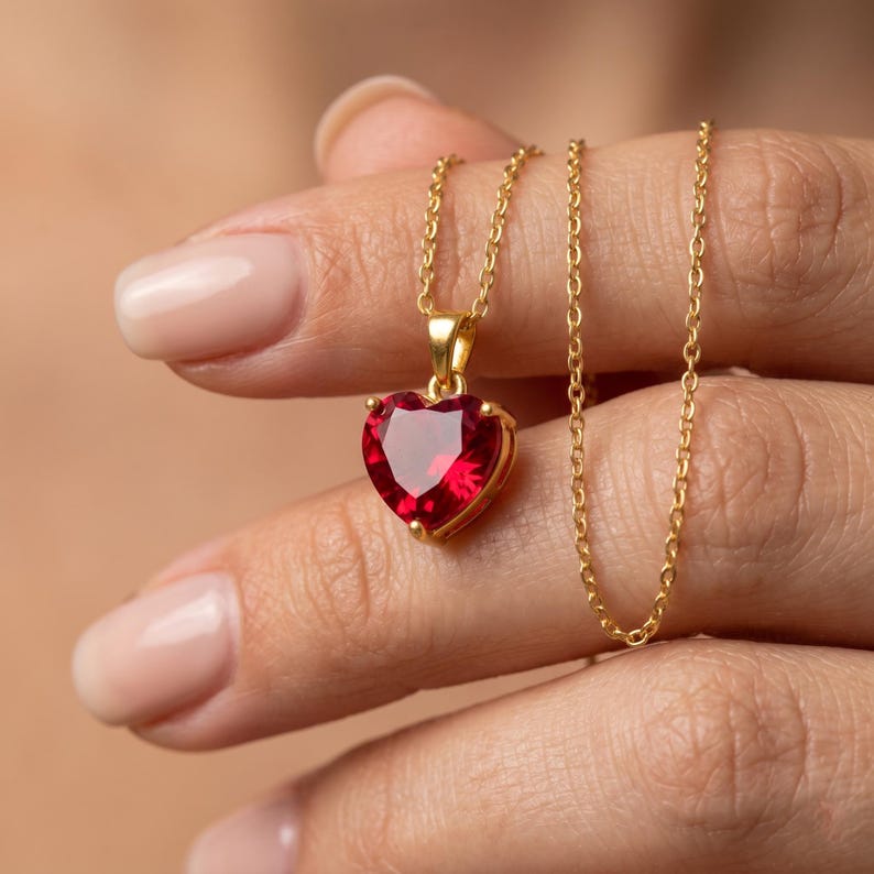 May include: A gold-colored necklace with a heart-shaped, ruby-red pendant. The pendant is a faceted gemstone set in a gold bezel. The chain is delicate and hangs from a gold bail.